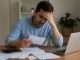 A person reviewing finances at a desk with bills, receipts, and a laptop, representing harmful money habits.
