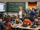 A German classroom scene with students and a teacher using digital and vocational tools, reflecting ongoing education system challenges.
